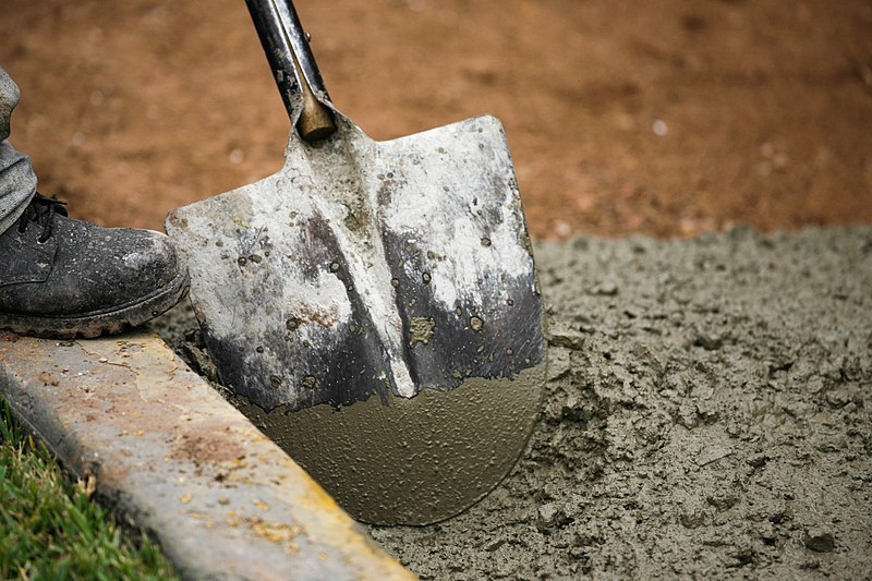 A shovel in a pool of wet cement; from the left of the frame, we see the toe of a work-boot, seemingly poised to plunge the shovel into the wet concrete.